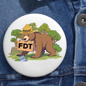 May include: A white button with a cartoon illustration of a brown bear wearing a yellow ranger hat with the word "RESIST" on it. The bear is holding a sign that says "FDT" in front of a green forest background.