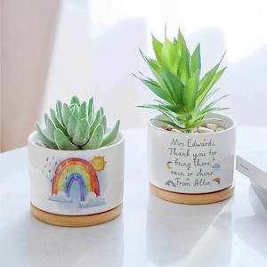 Puede incluir: Dos macetas de cerámica blanca con bases de madera, cada una con una suculenta. Una maceta presenta un diseño de arcoíris con un sol y lluvia. La otra maceta tiene el texto: "Sra. Edwards, Gracias por estar ahí, llueva o haga sol. De Alfie."