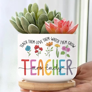May include: White ceramic planter with the text "TEACH THEM-LOVE THEM-WATCH THEM GROW TEACHER Mrs. Emily" and colorful flowers. The planter holds a succulent plant with green and red leaves, and sits on a wooden saucer.