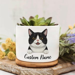 May include: White ceramic plant pot with a black and white cat illustration and the text "Custom Name". The pot contains a green succulent and rests on a wooden coaster. A delightful and personalized gift idea.