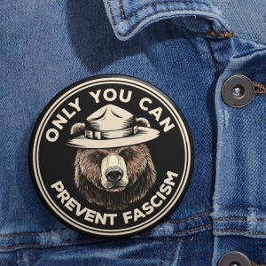 May include: A black and white button with a cartoon bear wearing a ranger hat. The text on the button reads "Only You Can Prevent Fascism."