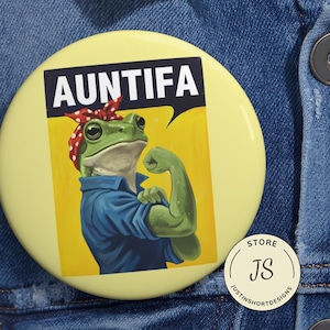 May include: A round, light yellow button with the word "AUNTIFA" in bold white letters. The button features a cartoon frog in a blue shirt and red bandana, flexing its arm. A small, white circular sticker with "STORE JS" is in the lower right.