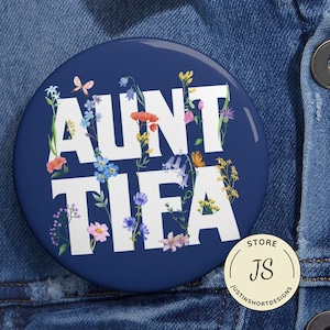 May include: Aunt Tifa button with a navy blue background. The white text is surrounded by colorful flowers and a butterfly. A small, round, gold-colored store logo is in the lower right corner.