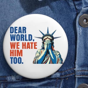 May include: White button with the text "DEAR WORLD, WE HATE HIM TOO." in blue and red. The Statue of Liberty is depicted with her hands covering her face in blue, red, and white.