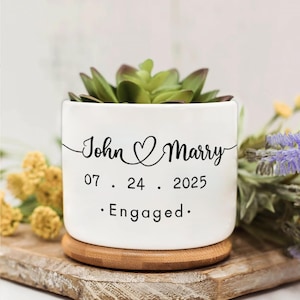 以下が含まれることがあります： 「John & Marry」の文字と日付「07.24.2025」、そして「Engaged」の文字が黒い筆記体で書かれた白いセラミックプランター。プランターには緑色の多肉植物が入っており、木製のコースターの上に置かれています。