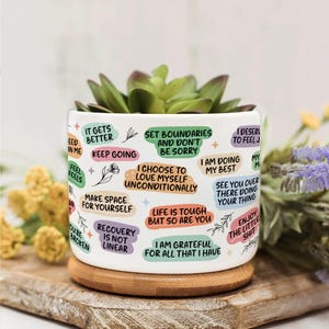 May include: White ceramic plant pot with a succulent plant. The pot is decorated with colorful text bubbles containing inspirational quotes. It sits on a round wooden coaster, with a wooden board in the background.
