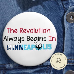 Może przedstawiać: Biały guzik z napisem "The Revolution Always Begins In MINNEAPOLIS" w kolorze czerwonym, czarnym i niebieskim. Guzik zawiera również symbol feniksa i małe logo sklepu.
