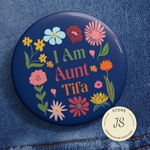 May include: A navy blue button with the text "I Am Aunt Tifa" surrounded by colorful flowers. The button has a glossy finish and is set against a denim background. A small, round store logo is in the lower right corner.