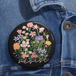 Puede incluir: Un botón negro con un diseño floral y la palabra "resist" en escritura rosa. Las flores son rosas, moradas y amarillas.