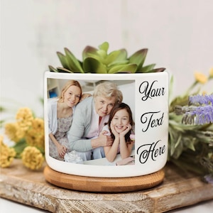 Puede incluir: Maceta de cerámica blanca con una foto familiar y el texto "Your Text Here". La maceta contiene una planta suculenta verde y se apoya en una base de madera. Un regalo personalizado.