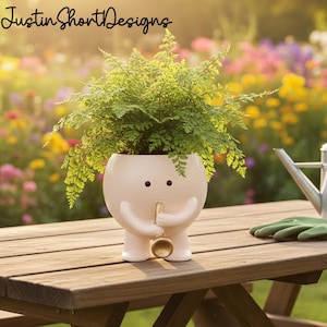 May include: A whimsical, light pink ceramic planter shaped like a character with arms, legs, and a trumpet. The planter holds a lush green fern. The planter is on a wooden table, with a blurred garden background. The text "Justin Short Designs" is at the top.