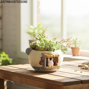 May include: A whimsical, off-white ceramic planter with a small house design, featuring a bunny figurine and a small red mushroom. The planter is filled with various green succulents and sits on a wooden table. The text "JUSTINSHORTDESIGNS" is visible.