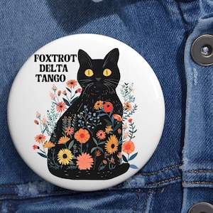 May include: A black cat with yellow eyes is surrounded by colorful flowers. The text "FOXTROT DELTA TANGO" is printed in black letters on a white background.