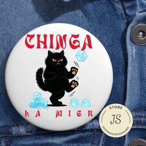 以下が含まれることがあります： 黒い猫が氷を蹴っているイラストが描かれた白いボタン。テキスト「CHINGA LA MIEB」は赤い様式化された文字です。小さなストアロゴが右下にあります。ボタンはデニムの背景にあります。