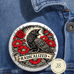 Może przedstawiać: Okrągła biała przypinka z czarną ilustracją kruka, czerwonymi różami i napisem "RADICALIZED By Basic Decency". Kruk otoczony jest ciernistym wieńcem i czerwonymi kwiatami. Małe logo sklepu widoczne jest w prawym dolnym rogu.