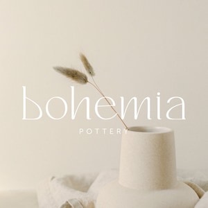 Könnte beinhalten: Eine weiße Keramikvase mit breiter Basis und schmalem Hals, die einen einzelnen Zweig getrockneten, beigen Pampasgrases hält. Die Vase steht auf einem beigefarbenen Leinenstoff. Der Text "bohemia pottery" ist in weißer Schrift unter der Vase geschrieben.