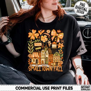 Pode incluir: Uma t-shirt preta com um gráfico colorido de uma cena de cidade pequena no outono. O design inclui edifícios, uma torre de água, flores, um laço e uma placa com o número 19008, com o texto SMALL TOWN FALL impresso na parte inferior.