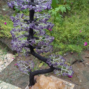 May include: A handcrafted bonsai tree sculpture with a black trunk and branches, adorned with purple crystal leaves. The tree is mounted on a light brown crystal base, set on a square stone tile. The background features green foliage.