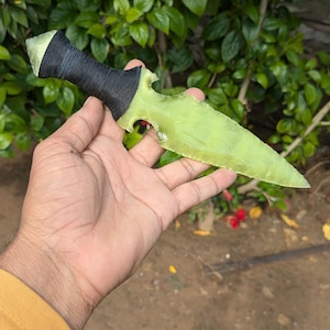 May include: A handmade, light green dagger with a black wrapped handle. The blade has a sharp point and a unique, asymmetrical shape. The dagger is held in a hand against a backdrop of green foliage.
