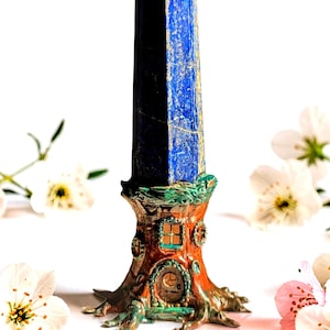 May include: A tall, blue lapis lazuli crystal obelisk, set on a whimsical, tree-trunk-shaped base with a miniature door and window. The base is painted in shades of brown, green, and orange. White flowers are scattered around the base.