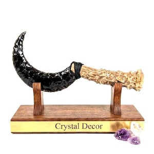 May include: A curved black obsidian blade attached to a textured tan antler handle with black cord wrapping. The object rests on a wooden stand with a gold-colored plaque reading Crystal Decor, accompanied by small purple and clear crystal pieces.