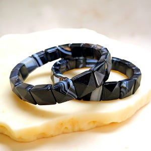 May include: Two black and white gemstone bracelets with geometric, faceted beads. The bracelets are displayed on a light-coloured, textured surface. The gemstones have a polished, reflective surface.