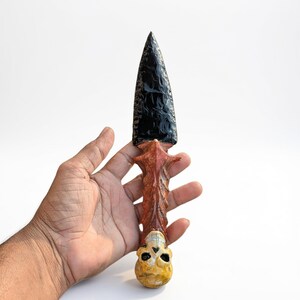 May include: A decorative dagger with a black, obsidian-like blade and a carved handle. The handle features a red and brown design with a skull at the base. The dagger is held in a hand against a white background.