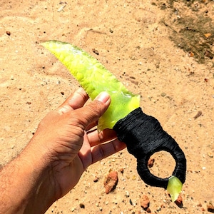 May include: A handmade knife with a translucent, lime-green blade and a black, wrapped handle. The handle features a finger loop. The knife is held against a backdrop of sandy ground.
