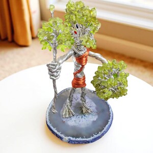 Könnte beinhalten: Eine handgefertigte Drahtskulptur einer Figur mit baumartigem Kopf und Armen, die einen Stab und eine Ansammlung grüner Perlen hält. Der Torso der Figur ist mit Kupferdraht umwickelt und steht auf einer blauen und grauen Achatscheibe.