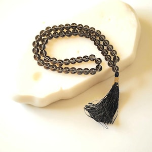 Op de afbeelding: Een ketting van rookkwarts kralen, gerangschikt in een gebogen patroon, met een zwarte kwast. De kralen zijn doorschijnend bruin van kleur. De kwast is bevestigd met goudkleurig draad. De kralen liggen op een wit, getextureerd oppervlak.