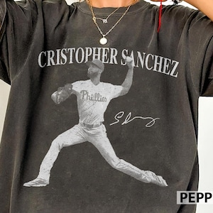 May include: Dark grey t-shirt with a black and white image of a baseball player. The text "CRISTOPHER SANCHEZ" is above the image, with "Phillies" on the jersey and a signature below. The shirt has a vintage, worn appearance.