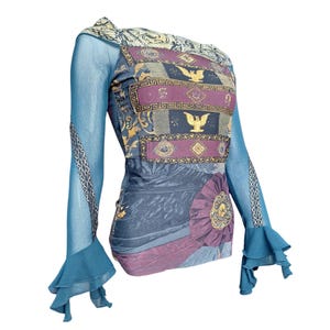 Peut inclure: Haut à manches longues avec un design unique. Le haut présente une combinaison de couleurs bleu, violet et or. Les manches sont bleu transparent avec des poignets à volants. Le corsage a un motif avec des accents dorés et une rosette décorative.