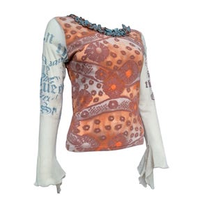 May include: A long-sleeved top with a unique design. The body of the shirt is orange with a floral pattern, while the sleeves are off-white with blue text. The neckline is adorned with a blue trim, and the sleeves flare at the wrists.