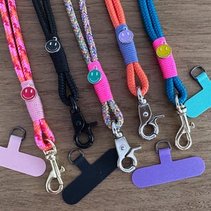 Könnte beinhalten: Fünf bunte Seil-Lanyards mit drehbaren Metallkarabinerhaken und kleinen Smiley-Anhängern. Jedes Lanyard hat ein anderes Kordelmuster, darunter Pink-Orange, einfarbig Schwarz, Mehrfarbig, Orange und Blau, ergänzt durch passende Handy-Tabs.