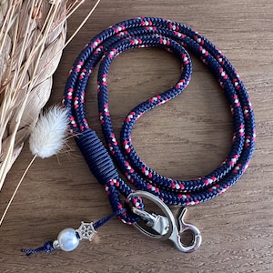 Könnte beinhalten: Ein marineblaues Schlüsselband mit roten und weißen Akzenten. Das Schlüsselband besteht aus einem dicken, seilartigen Material und verfügt über einen silberfarbenen Verschluss und einen dekorativen Anhänger mit einer Perle. Das Schlüsselband wird auf einer Holzoberfläche präsentiert.