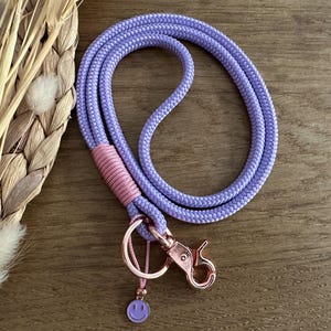 Könnte beinhalten: Ein lilafarbenes Schlüsselband mit einem rosa Akzent und einem roségoldenen Verschluss. Das Schlüsselband hat einen Smiley-Anhänger und besteht aus einem dicken, gewebten Kordel. Der Hintergrund ist eine Holzoberfläche.