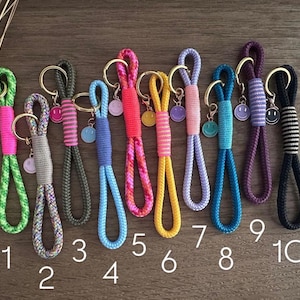 Könnte beinhalten: Zehn farbenfrohe Schlüsselanhänger aus Seil in einer Reihe, jeder mit einem goldfarbenen Metallring und einem kleinen baumelnden Smiley-Anhänger. Die Schlüsselanhänger variieren in der Farbe, darunter Neongrün, Grau, Oliv, Blau, Pink und Schwarz.