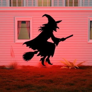 May include: A black silhouette of a witch riding a broomstick, set against a pink-toned house exterior. The witch is wearing a pointed hat and flowing robes. The scene is illuminated with a red glow, suggesting a Halloween theme.