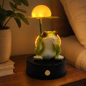Könnte beinhalten: Eine grüne Frosch-Figur-Lampe mit roten Augen, die eine gelbe, pilzförmige Leuchte über dem Kopf hält. Der Frosch sitzt auf einem runden schwarzen Sockel mit einem goldfarbenen Knopf an der Vorderseite und strahlt ein warmes Licht aus.
