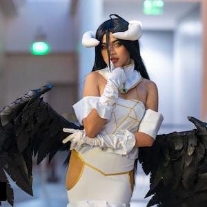 May include: A woman in a white and gold costume with black feathered wings. She is wearing a white headband with horns and has long black hair. She is posing with her hand on her chin.