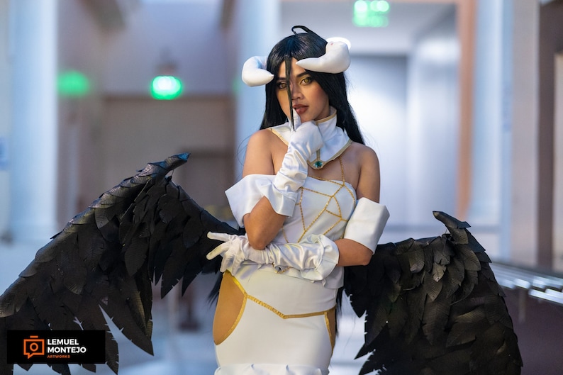May include: A woman in a white and gold costume with black feathered wings. She is wearing a white headband with horns and has long black hair. She is posing with her hand on her chin.