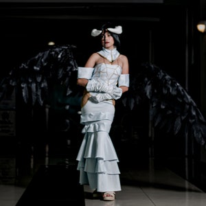 May include: A woman in a white dress with black feathered wings. She is wearing a white headband with a gold design and has a serious expression on her face.