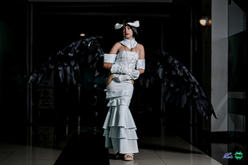 May include: A woman in a white dress with black feathered wings. She is wearing a white headband with a gold design and has a serious expression on her face.