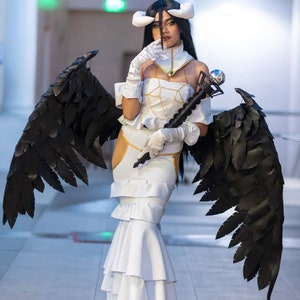 May include: A woman in a white dress with black angel wings holds a staff with a silver orb on top. She is wearing a white headband with horns and a gold necklace. The background is a white hallway.