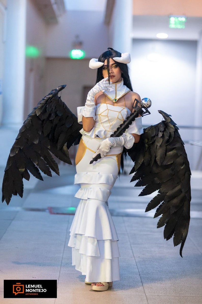 May include: A woman in a white dress with black angel wings holds a staff with a silver orb on top. She is wearing a white headband with horns and a gold necklace. The background is a white hallway.