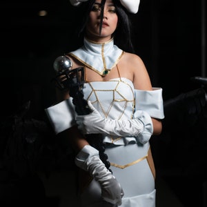 May include: A woman in a white and gold costume with a black and silver staff. She is wearing white gloves and has horns on her head. The background is dark.