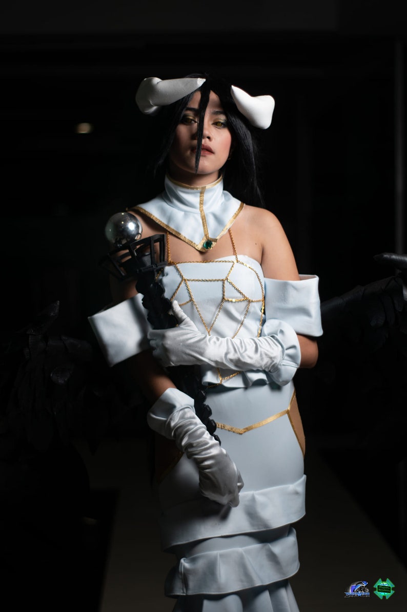 May include: A woman in a white and gold costume with a black and silver staff. She is wearing white gloves and has horns on her head. The background is dark.