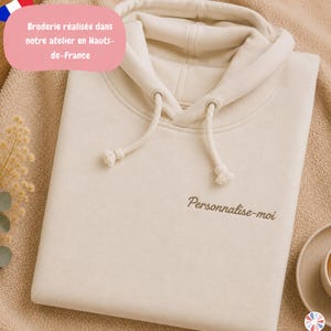 Peut inclure: Un sweat à capuche de couleur crème doté d'une capuche à cordon de serrage avec des cordons assortis et des œillets métalliques. La poitrine présente le texte brodé 'Personnalise-moi' dans une police cursive marron foncé centrée.