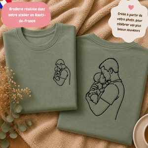 Peut inclure: Deux t-shirts vert sauge ornés d'une broderie au trait noir représentant une personne tenant un bébé. Les t-shirts sont posés sur un tissu beige texturé. La broderie montre le profil d'un adulte berçant un nourrisson dans ses bras.