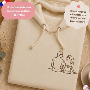 Pode incluir: Uma camisola com capuz de cor creme com um bordado de linha castanho-escuro de duas pessoas sentadas lado a lado. A camisola inclui um cordão a condizer com ilhós metálicos e um tecido macio com uma textura visível.
